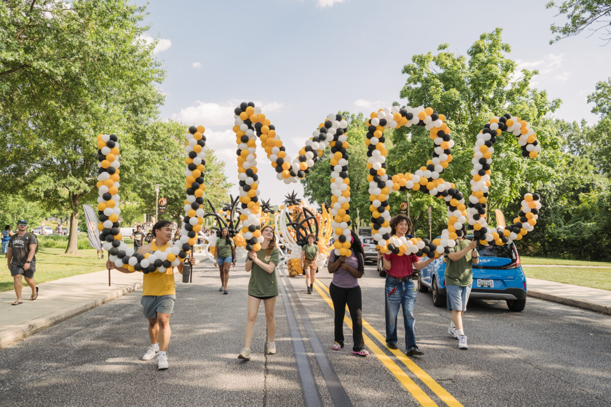 students parade down the street holding large balloons that spell out UMBC