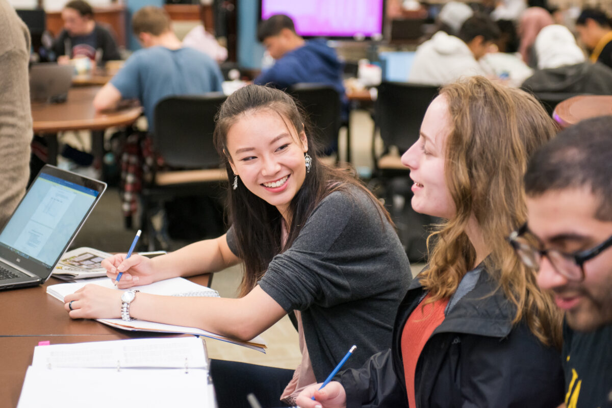 two students sit at a table, study materials laid out in front of them, one smiles at the other; many other students seated in groups at tables in the background