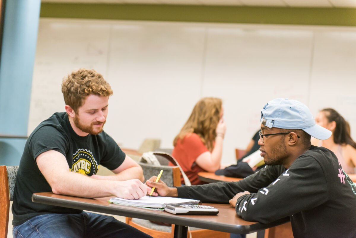 two students sit across from each other at a table, both looking at a notebook between them, one is pointing to it with a pencil, whiteboards in the background 