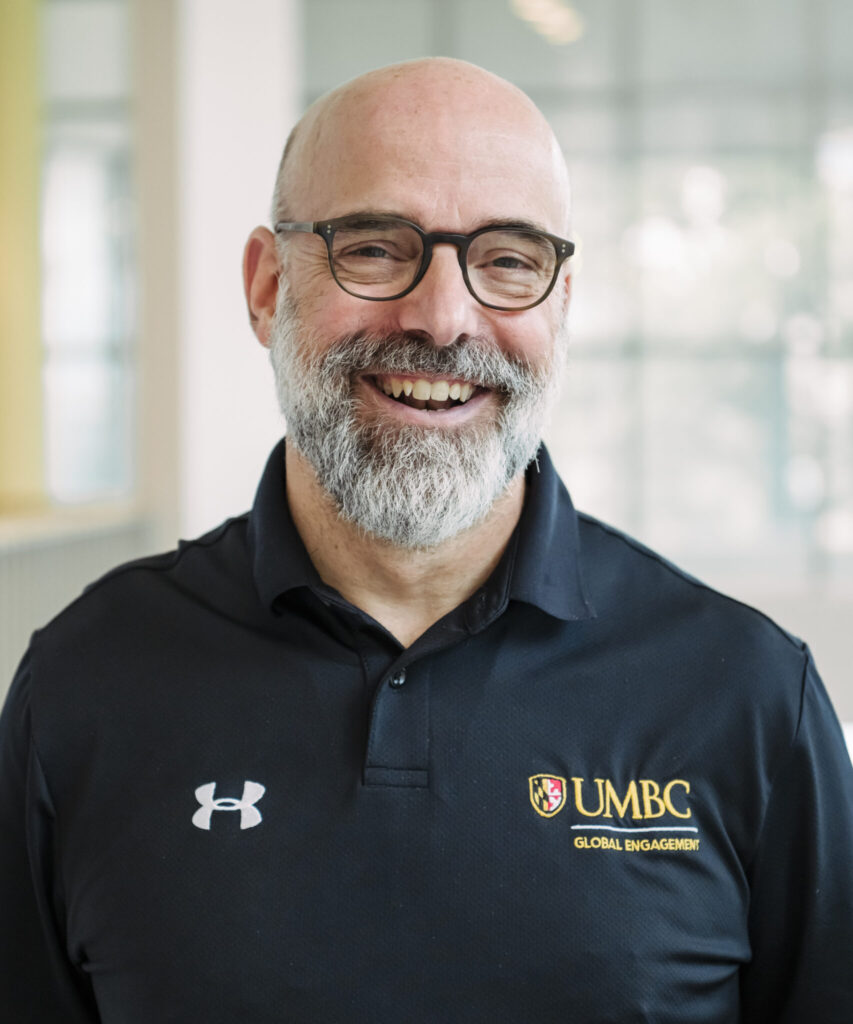 An adult with a bald head and grey beard wearing a black polo shirt with the under armor  and UMBC logos 2024 – 2025 Fulbright