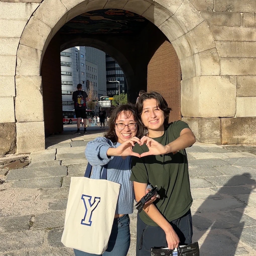 Two people stand close to each other holding their hands out to meet in the middle creating a heart shape with their fingers