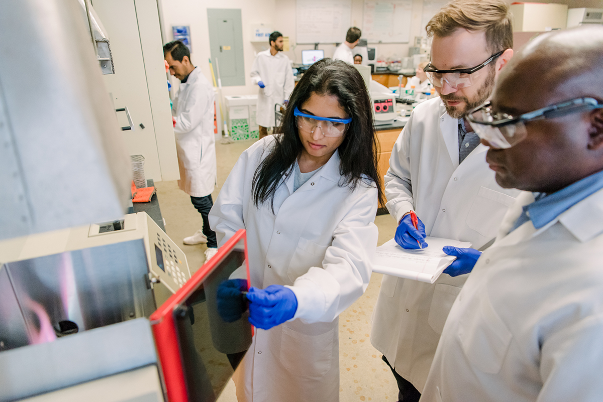 People wearing lab coats, gloves, and safety glasses stand in a lab. In the center, a woman opens the door of a piece of lab equipment while two men stand nearby, one holding a pen and paper.