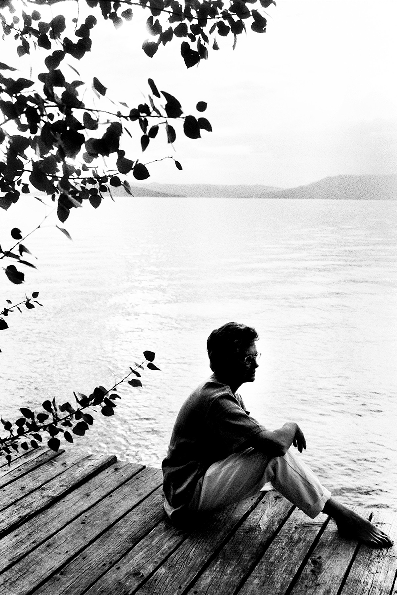 In a black and white photo, a person on a dock looks out across a river.