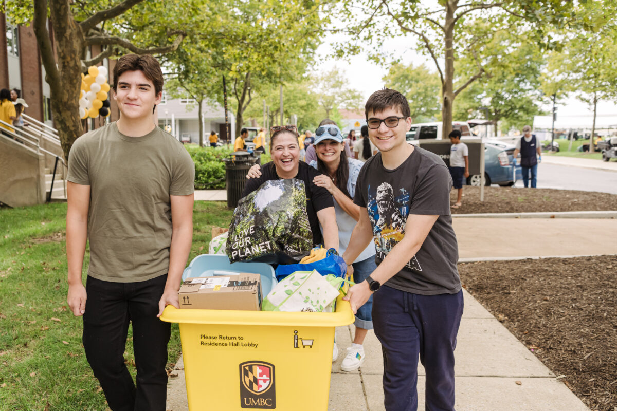 Two students push a cart with dorm supplies while two women smile behind them