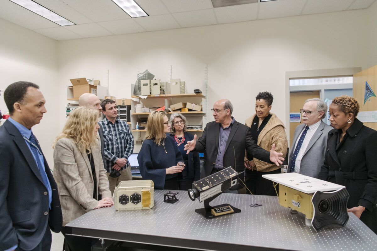 A room of people looking at the Hyper-Angular Rainbow Polarimeter instrument family. One person in the center has his hands outstretched, looking at someone on his left as he explains what the instruments do.