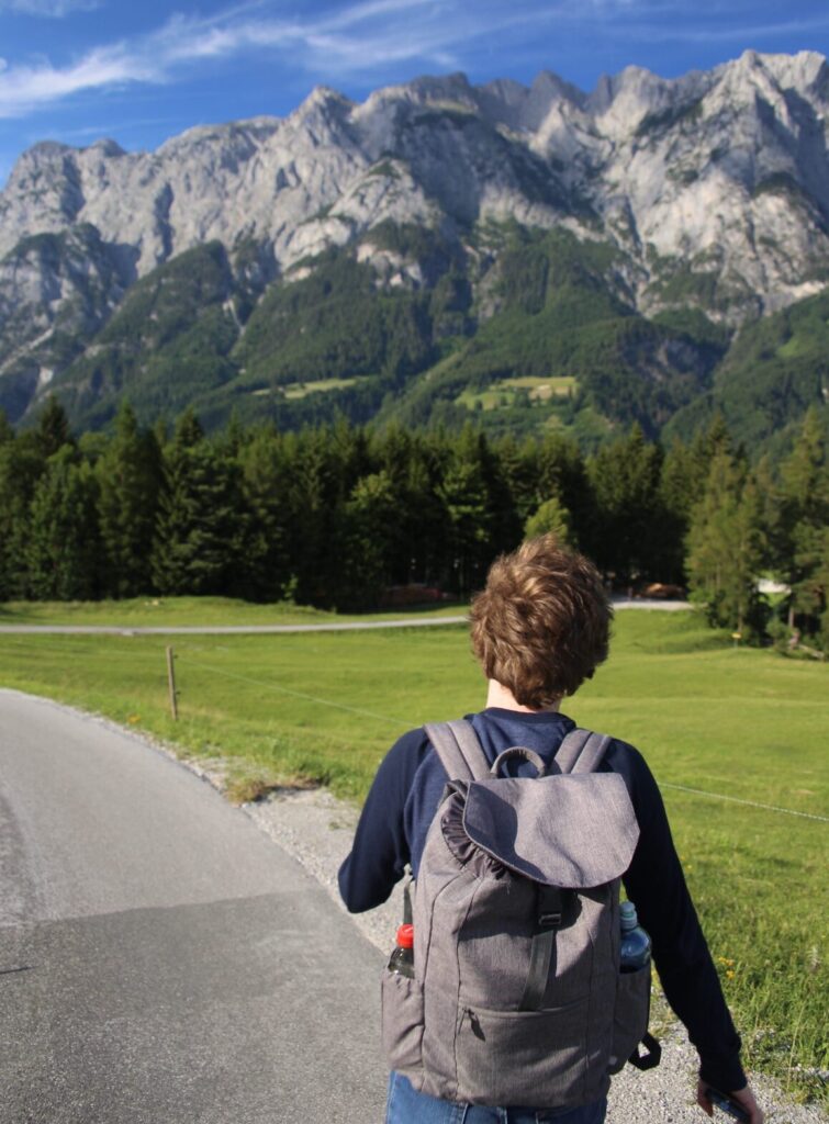 A college student with a grey backpack on their back walking on a paved road toward large mountains in Austria. Education abroad scholarship