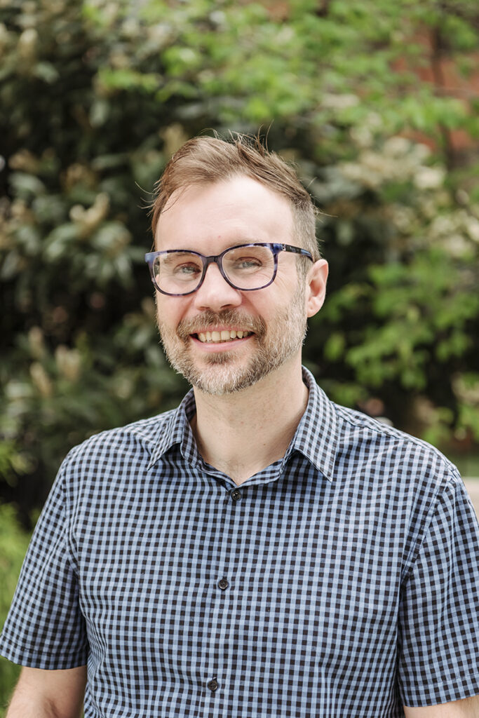 A man wearing glasses and checkered shirt smile at camera in front of greenery.