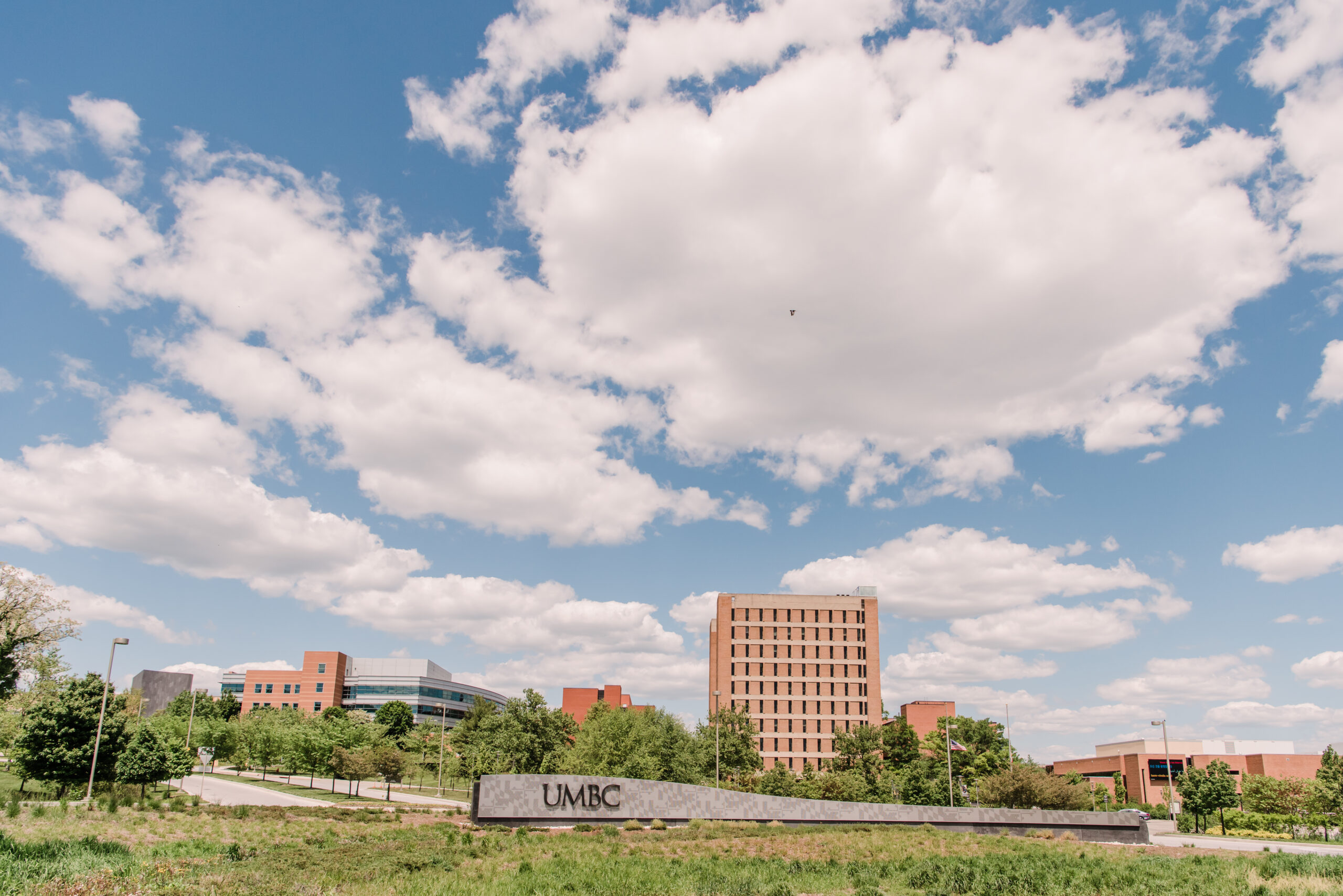 One of the entrances of UMBC's campus. There is a UMBC sign in front of a tall building. The sky is blue and clouds are in the sky.