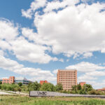 One of the entrances of UMBC's campus. There is a UMBC sign in front of a tall building. The sky is blue and clouds are in the sky.