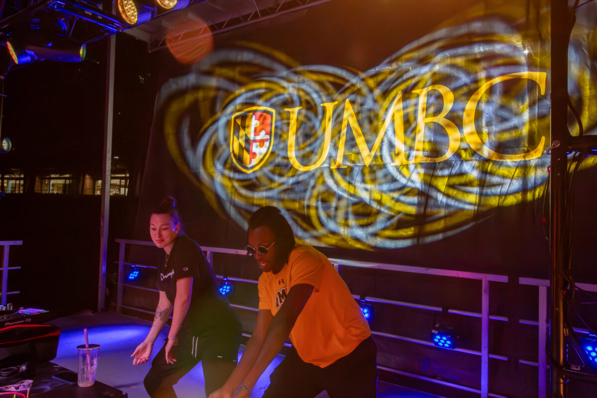 Two DJs stand at a table with UMBC projecte behind them