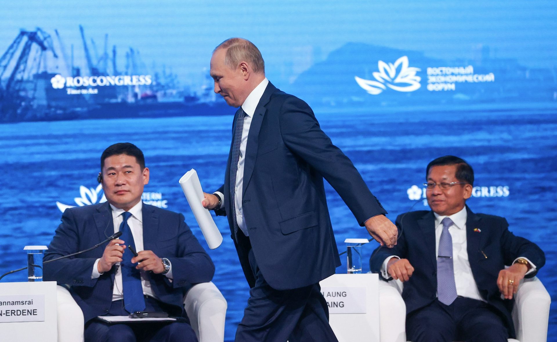 Vladimir Putin, president of Russia, walks across a stage in front of two men sitting on white chairs