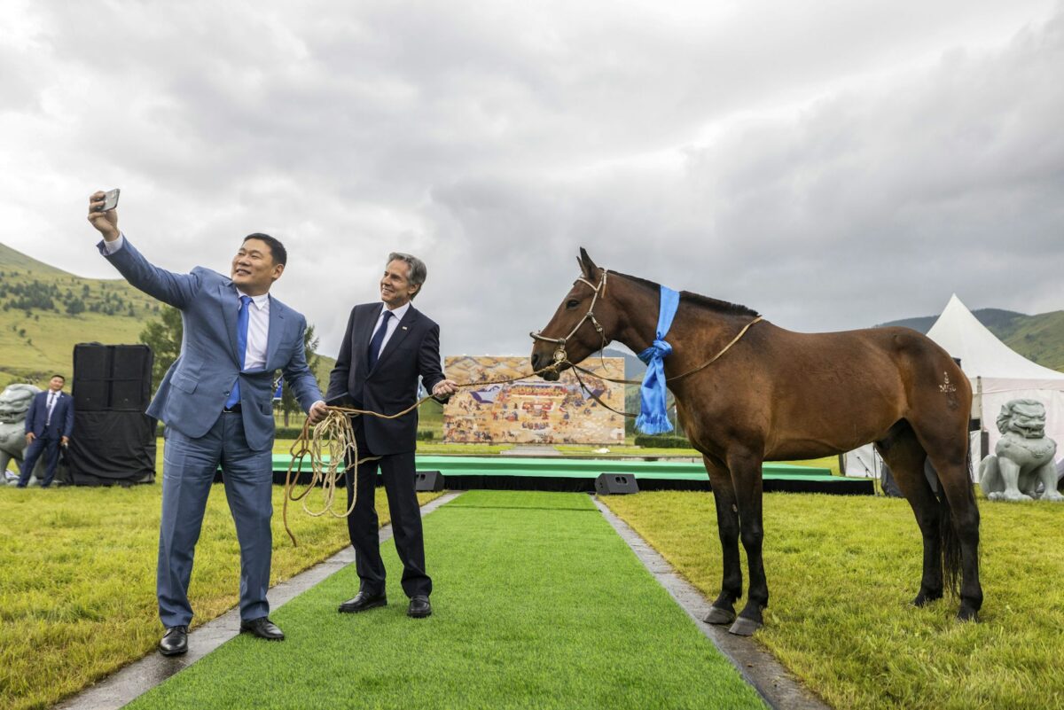 Two men in business suits take a selfie of themselves holding a horse's rains while standing on a green field in front of a stage.