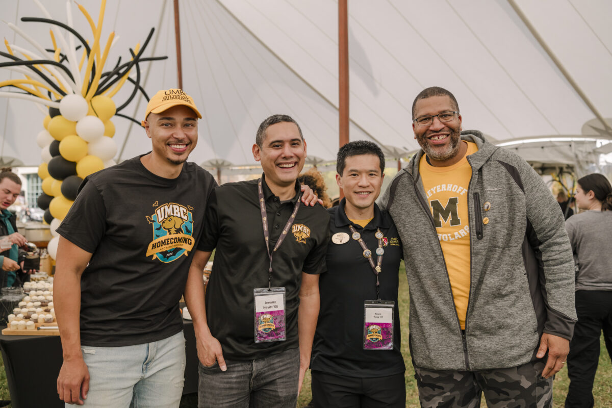 four men smiling at the camera. they are all wearing umbc apparel. there are black, yellow, and white balloons behind them 