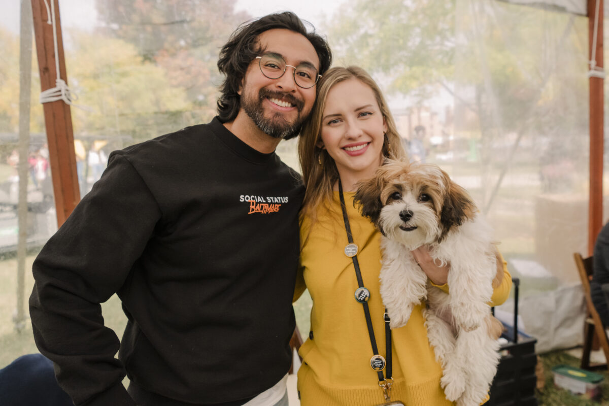 two people smiling at the camera. the person on the right is holding a mostly white dog with brown ears