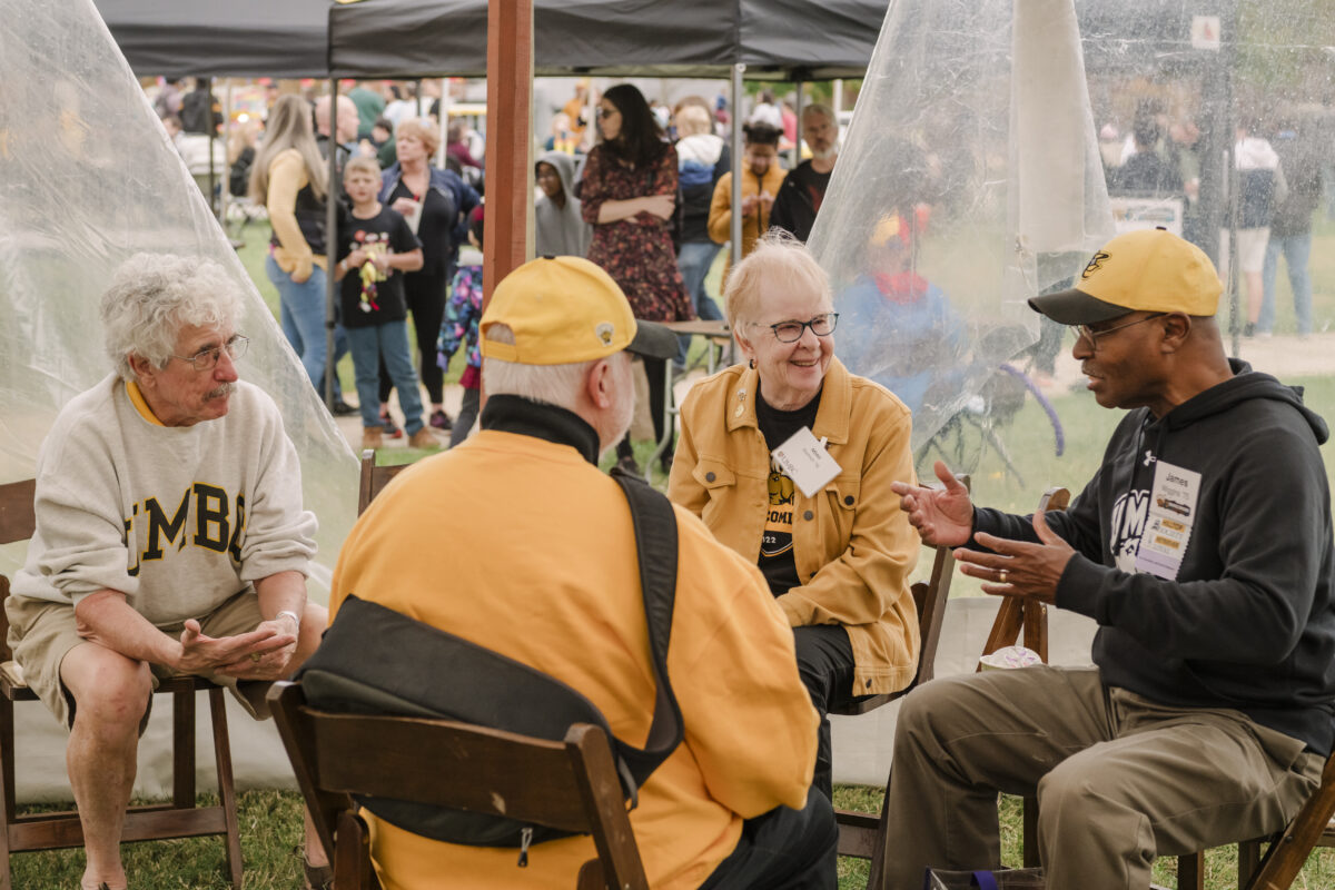 four older people sitting outside of a tent talking. they are sitting on chairs. one person on the right is gesturing with his hands.