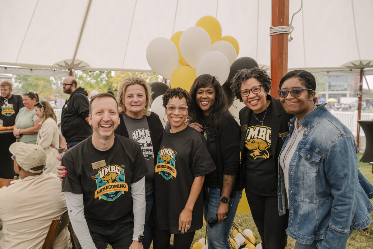 a group of six people smiling at the camera. there are yellow and white ballons in the baground 