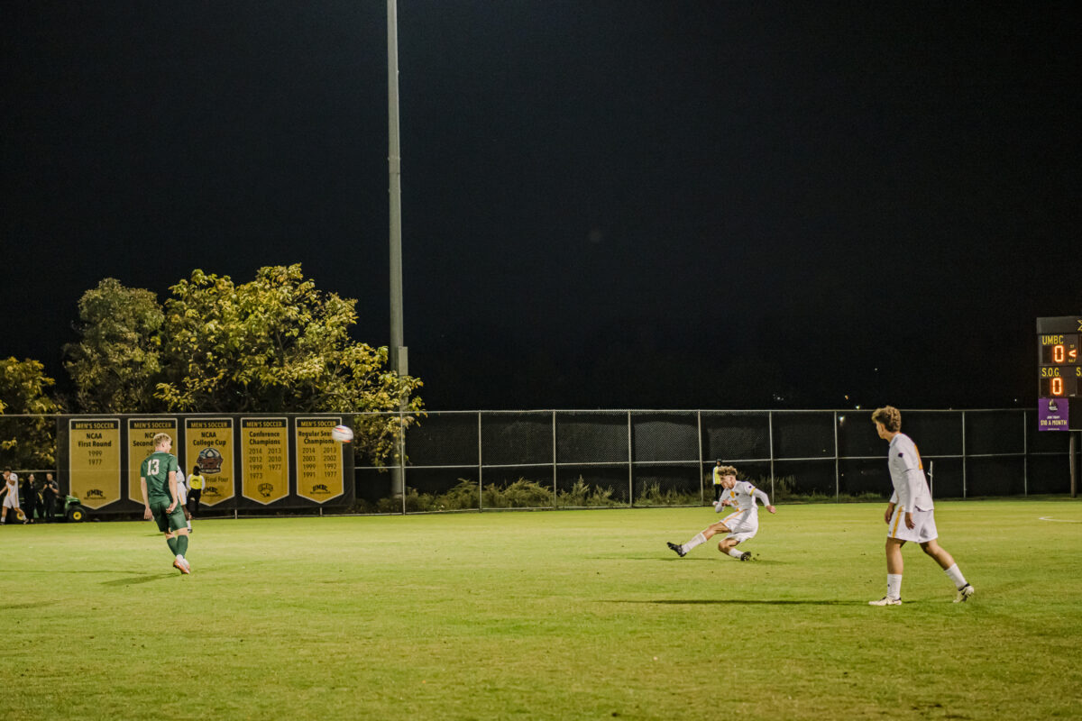 three soccer players on a field.  two on the left are in white UMBC uniforms and one on the left is in a  green uniform. one player is in the motion of kicking, the ball is flying in the air to towards the player in green. 