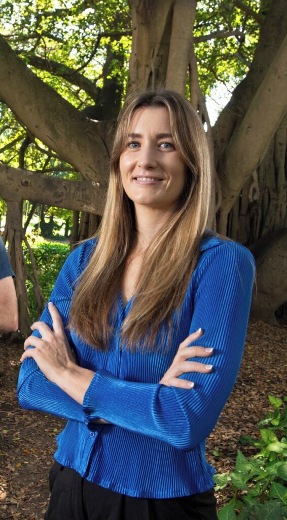 smiling woman in a bright blue top standing with her arms folded in front of a huge tree