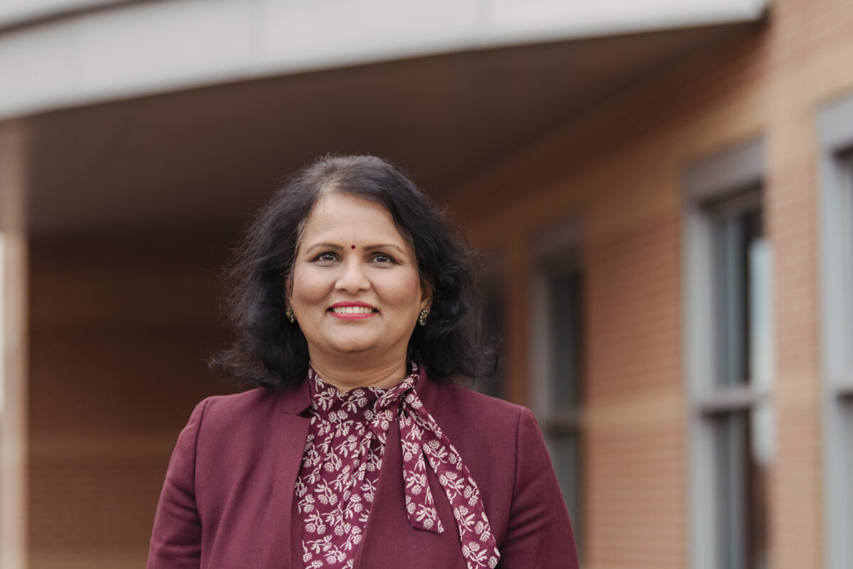 Woman is wearing a burgundy suit jacket and is smiling at the camera. 