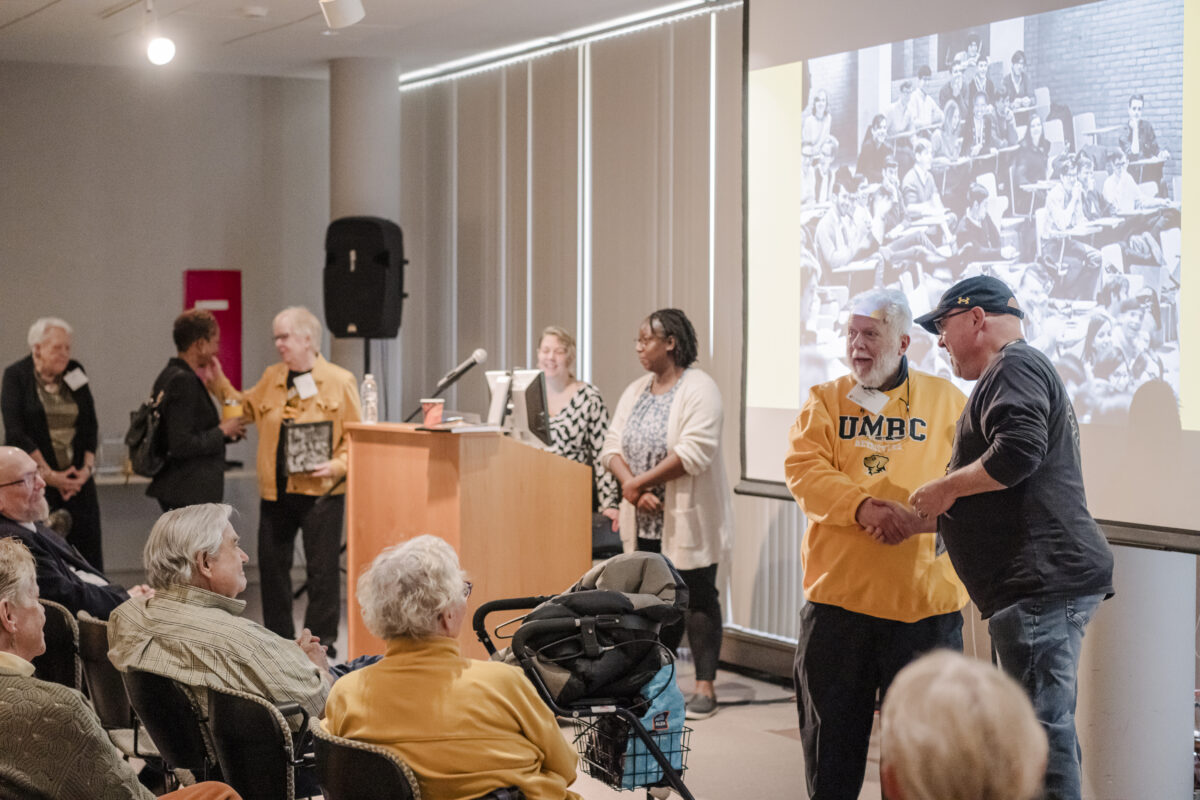 people in a room. one person is standing behind a podium there are two men on far right shaking hands. there are many people sitting in the audience. 