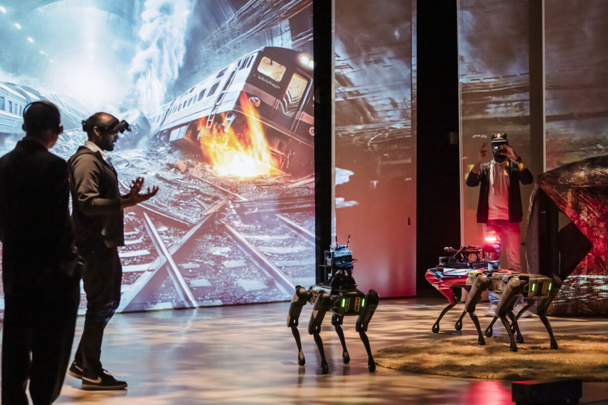 Researchers on stage with two autonomous robots that look like robot dogs. The researchers have on headgear that they are using the direct the robots. There is a picture of an animated train crash in a tunnel on the walls behind the researchers. 