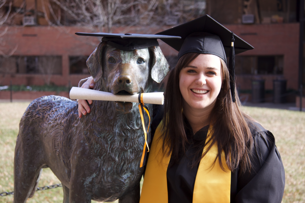 A woman in graduation regalia poses with a chesapeake bay retriever statue with a diploma in it's mouth.