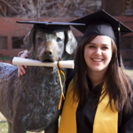 A woman in graduation regalia poses with a chesapeake bay retriever statue with a diploma in it's mouth.