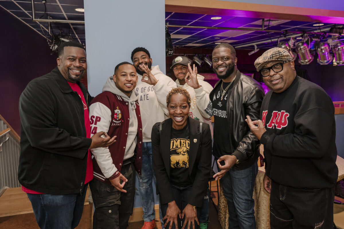 seven people smiling for the camera. there are 6 men who are holding up their fraternity's hand sign. one woman in the middle is crouching. 