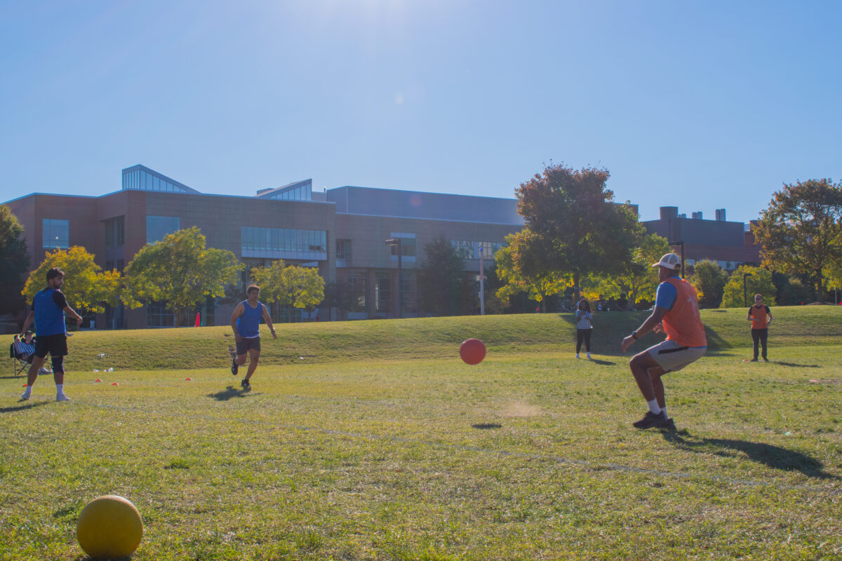 UMBC students on a field playing kickball. there is an orange kickball in  the air as one person begins to run.