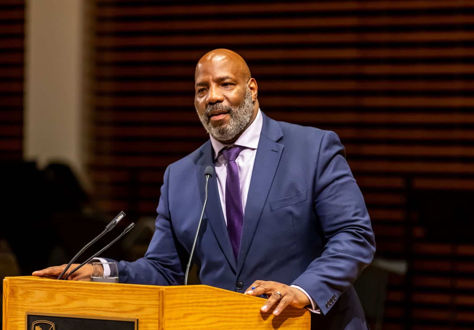 A person with a medium dark skin tone, wearing a blue jacket and purple tie, stands at a lectern.