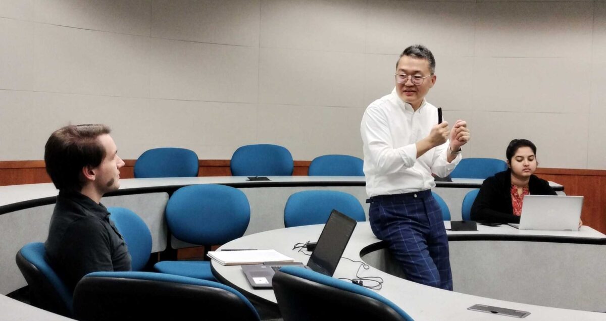 Man in glasses talks with students seated in circular rows of desks and chairs in lecture hall.