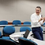 Man in glasses talks with students seated in circular rows of desks and chairs in lecture hall.