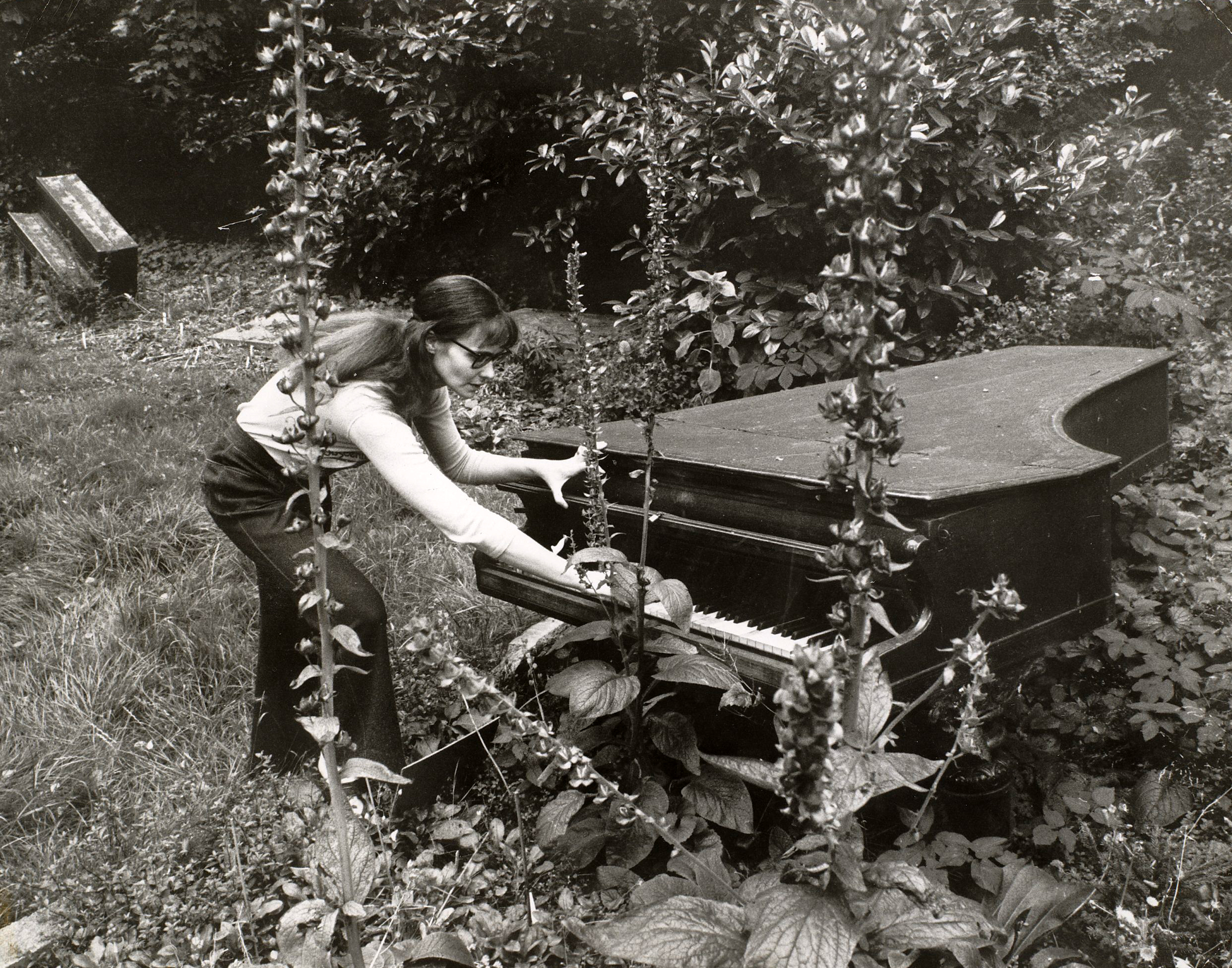 A person plays a piano that has been set outside and is covered with foliage.