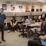 A group of college students sitting around rectangular desks. There is a person standing near the left hand side who is pointing up towards something out of camera.