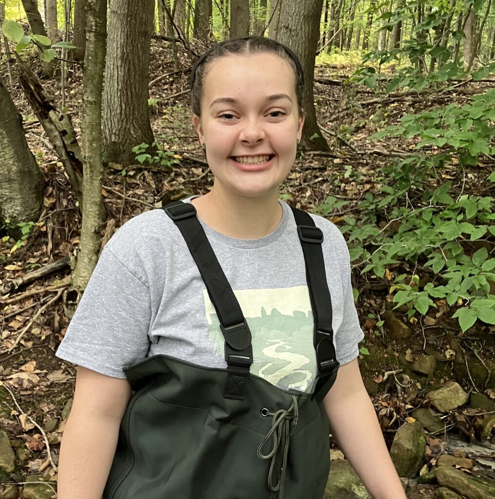 woman in a gray t-shirt and waist-high waders standing in a forest