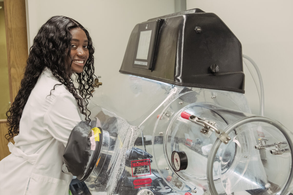 smiling student stands near a large clear enclosure in a laboratory used to control oxygen levels