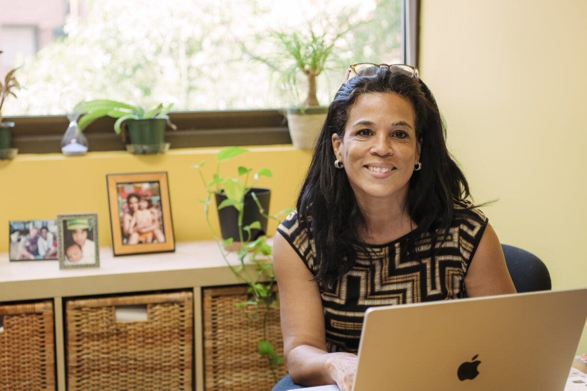 woman smiles out from sitting at a desk with a mac laptop; warm wood furnishings, a few plants, and family photos in the background