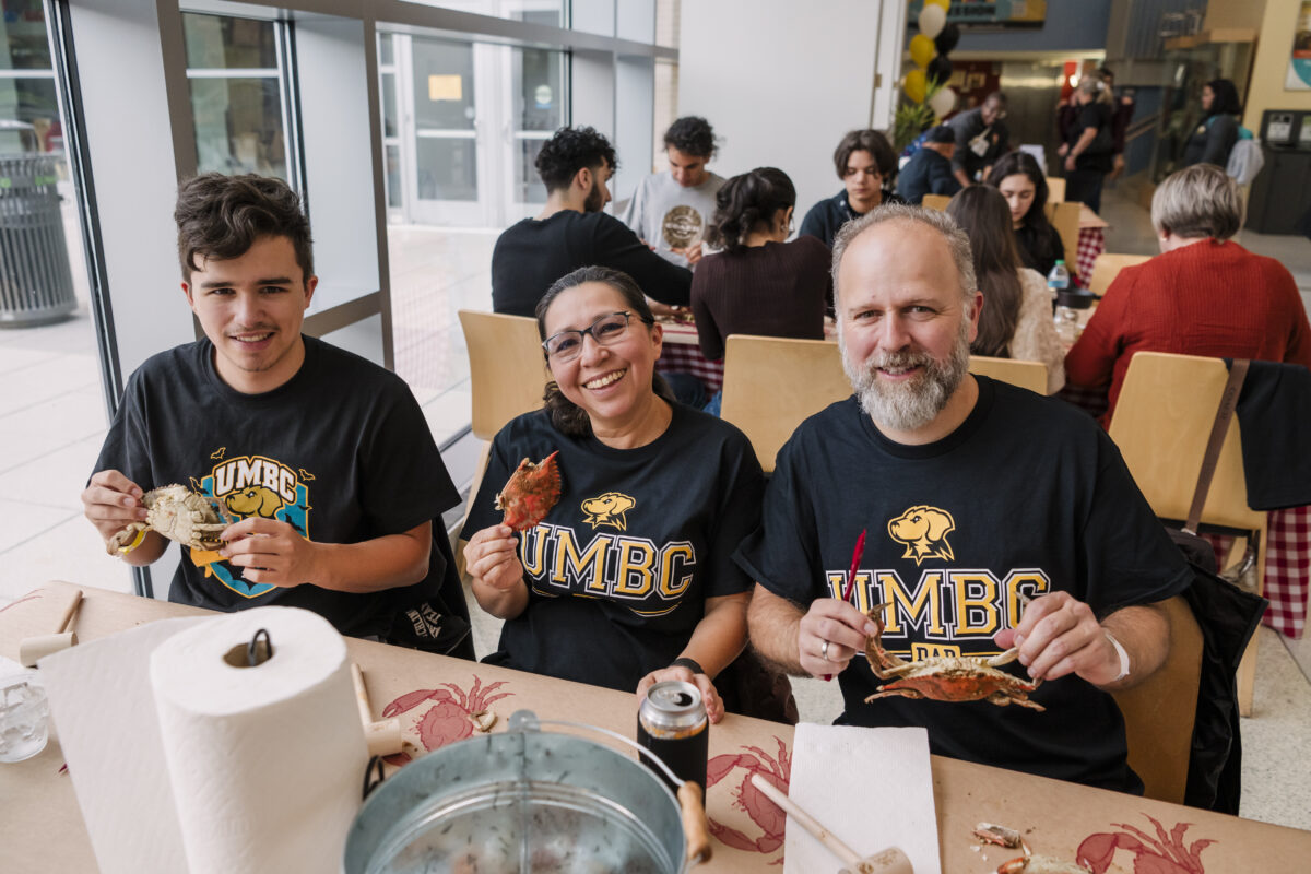 a family holding up crabs and smiling. they are sitting at a table that is covered with paper towel holder, a bucket for the crabs, and various crab cracking utensils. 