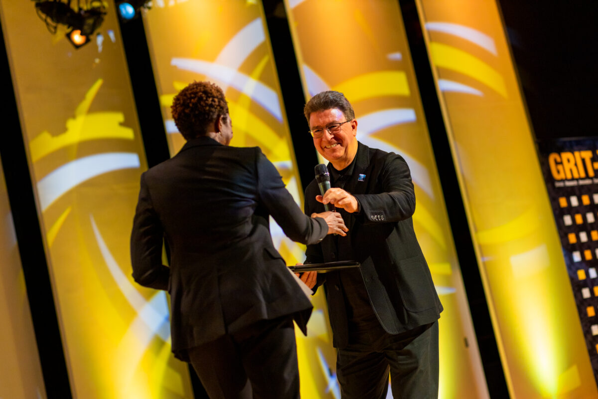 Two people on a stage, one of them has their back turned away from the camera. The man on the right is extending a microphone to woman on the left.