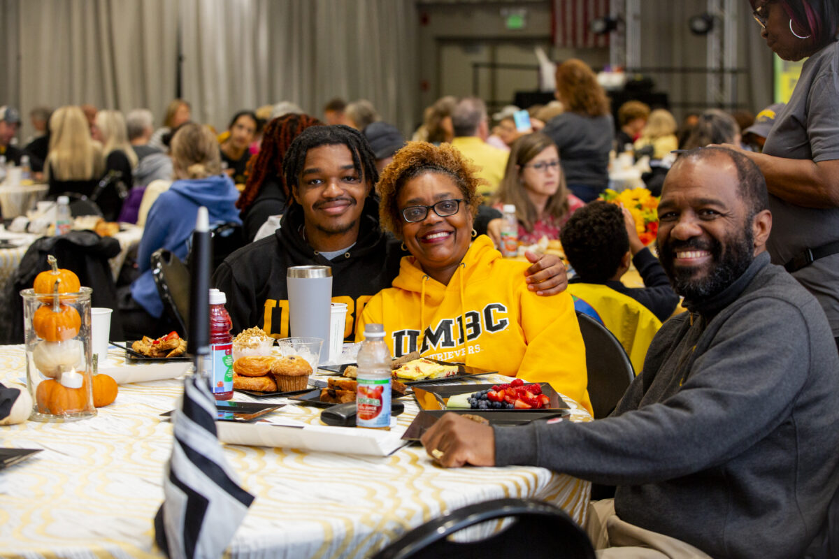 Family wearing UMBC sweatshirts sit at table with breakfast food. They smile at the camera.