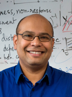 A man who is wearing rectangular glasses and a blue collared shirt is smiling at the camera. 