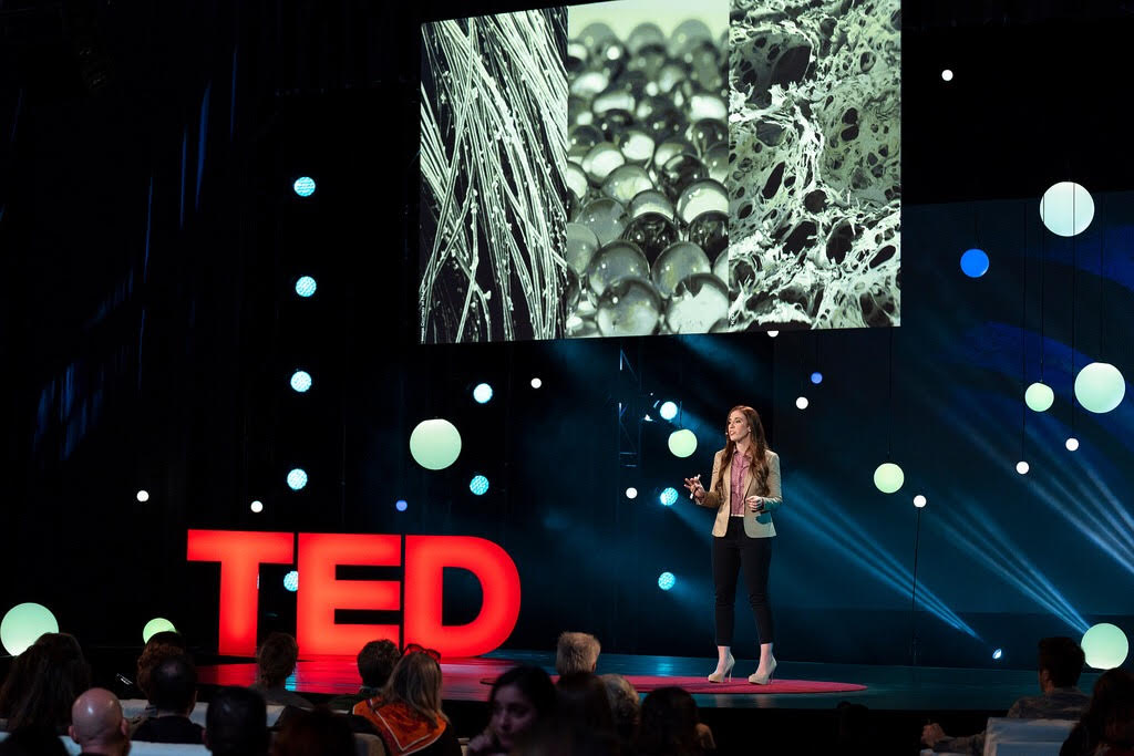 TIME100 Next list recipient Katilyn Sadtler stands on stage, large red "TED" sign to the left, screen showing what looks like a microscope image of cells behind her