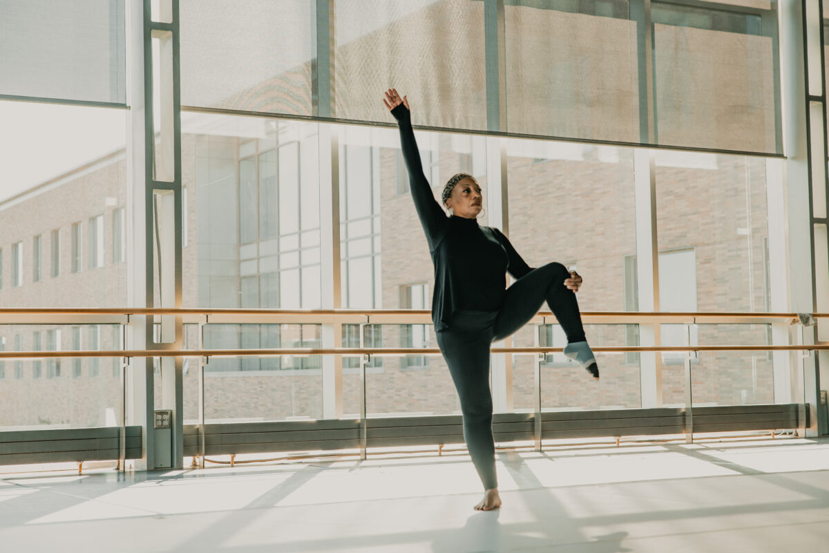 Teresa Kearney gracefully balances on one leg in a dance studio. One arm is extended upward and the other holds up her corresponding leg with toes pointed towards the floor. The large windows behind her bring in sunlight, with slight shadows of the windows on the dance floor