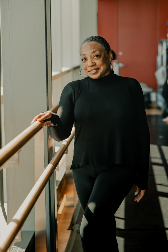 Wearing black dance attire, Teresa Kearney stands by the ballet barre in a dance studio. Her hand gently rests on the rail. She faces the camera and smiles warmly. Sunlight comes in through the large windows next to her.

