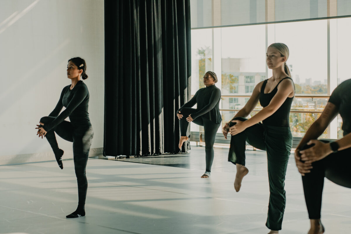 A group of dancers in black attire, including Teresa Kearney, balance on one leg in a bright dance studio. Each dancer holds their raised knee with both hands while maintaining poise and focus. 