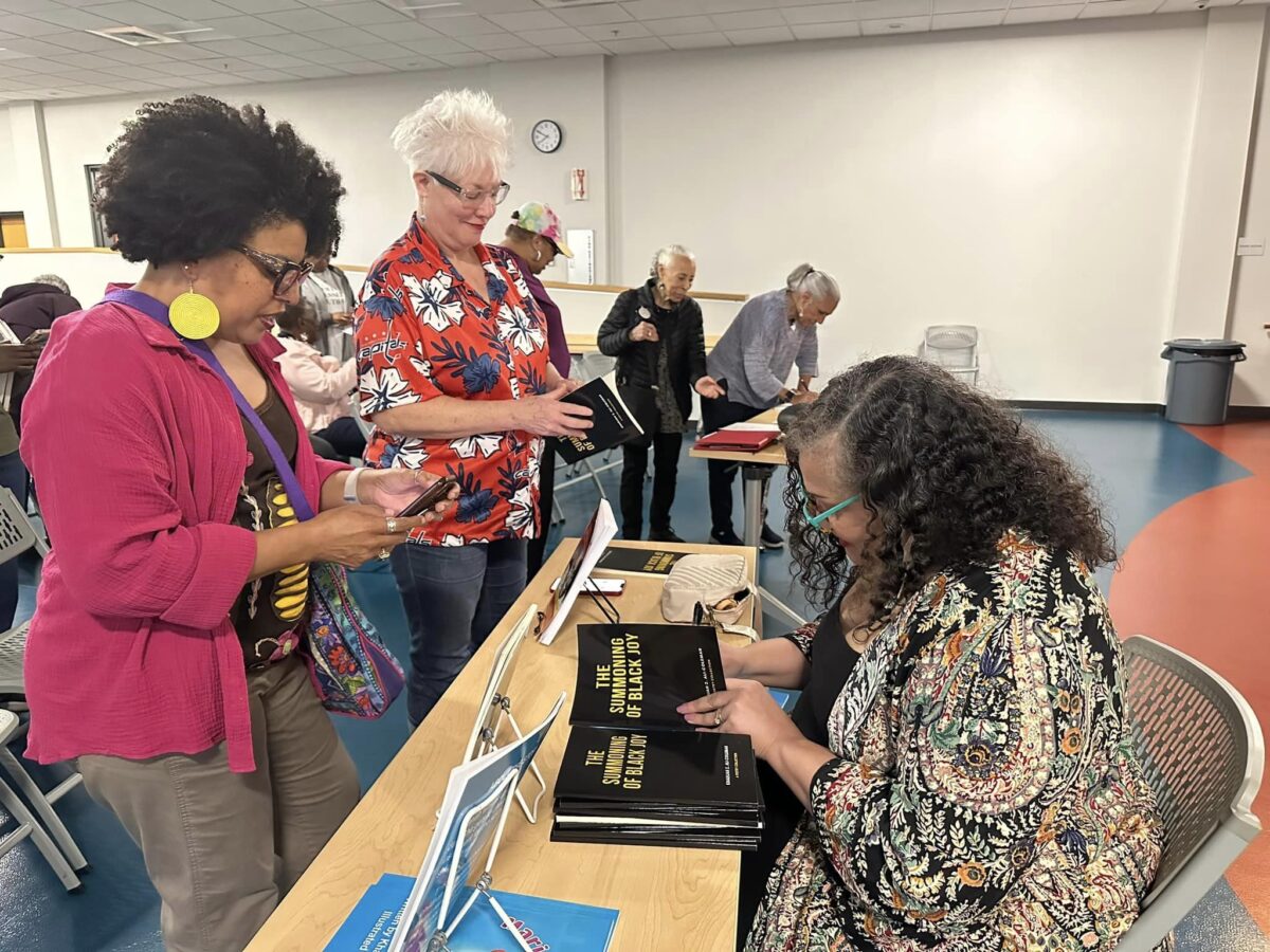 A woman signs a book at a table during a presentation for an audience member. 