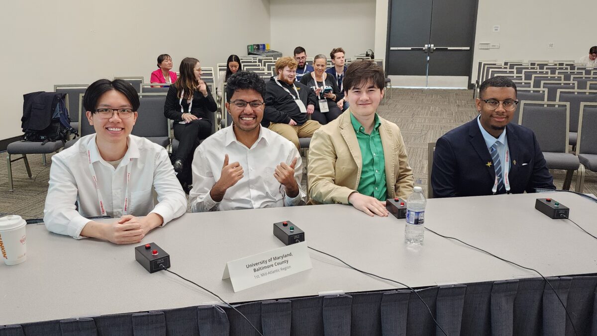 Four students sit behind buzzers at front table. Audience members sit behind.