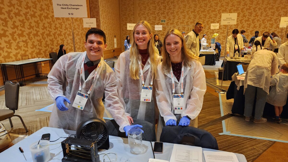 Three students in lab coats stand behind table with beaker, fan, and other equipment.