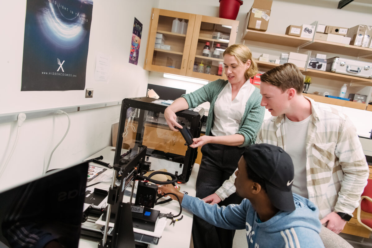 professor and two students work at a large table, handling various electronic and mechanical parts.