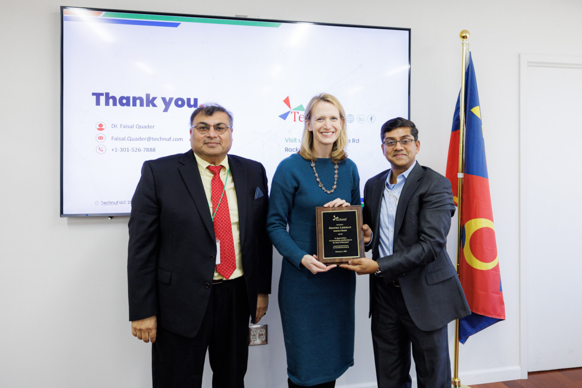 Three people posing for a picture while standing in front of a smart monitor that says words on it. We can see the words thank you on the screen. The woman in the center is holding a plague with writing on it. The man on the right is holding the plague with her.  